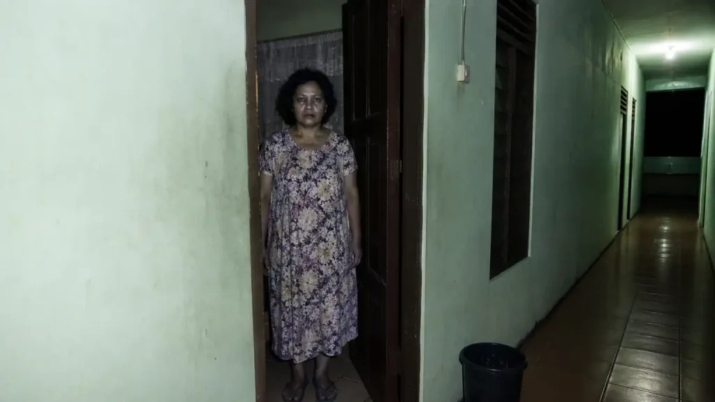 Visual realistis penghuni misterius berdiri di pintu kamar kos Indonesia pada malam hari